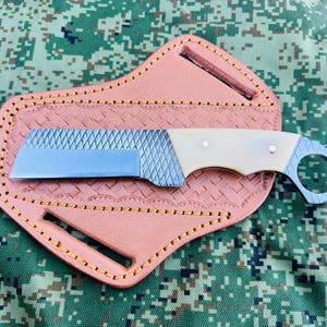 May include: A custom-made knife with a textured blade and a cream-colored handle, resting on a light brown leather sheath. The sheath has a decorative pattern and is stitched with gold thread. The knife has a ring at the end of the handle.