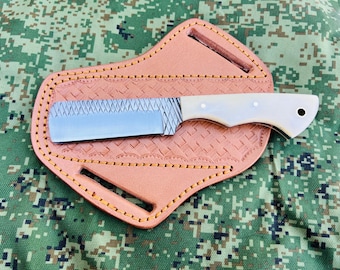 May include: A handmade knife with a textured blade and a white handle rests in a tan leather sheath. The sheath has decorative stitching and a cutout for a belt. The background is a camouflage pattern.