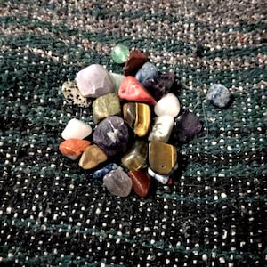 May include: A collection of polished gemstones in various colors, including purple, brown, yellow, red, and green. The stones are arranged on a textured, dark green and white speckled surface. The gemstones vary in size and shape.