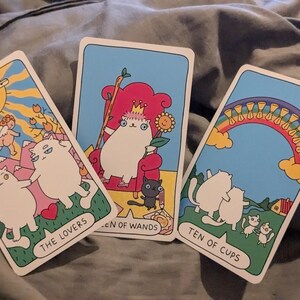 Könnte beinhalten: Drei Tarotkarten mit Cartoon-Katzen. Die linke Karte trägt den Titel The Lovers, die mittlere Queen of Wands und die rechte Ten of Cups. Jede Karte zeigt farbenfrohe Illustrationen von weißen Katzen in verschiedenen Szenen vor einem hellblauen Himmel.