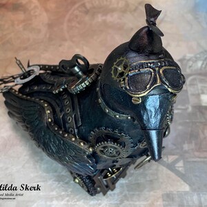 May include: A black steampunk bird figurine with gold accents and gears. The bird has a propeller on its head and goggles over its eyes.