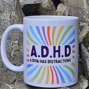 May include: A white ceramic mug featuring a colourful rainbow sunburst pattern radiating from the centre. The text A.D.H.D is printed in large purple letters, with the phrase A DIVA HAS DISTRACTIONS written in smaller black capital letters below it.