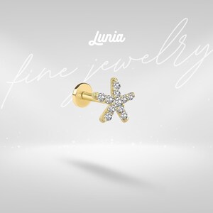 May include: A gold-toned flat back stud earring featuring a five-pointed star shape adorned with clear, sparkling stones. The text Lunia fine jewellery is visible in the background. The piece has a smooth, polished metal finish and a circular base.