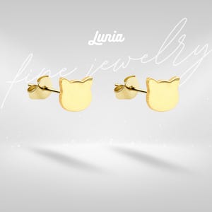 Op de afbeelding: Een paar goudkleurige katvormige oorstekers. De oorbellen hebben een gladde, gepolijste afwerking. De merknaam "Lunia" staat bovenaan de afbeelding, met de woorden "fine jewelry" in een cursief lettertype.