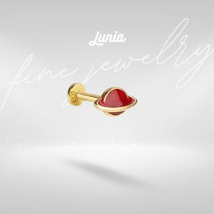 Puede incluir: Pendiente de botón dorado con un diseño de planeta rojo y un anillo dorado. El pendiente tiene una parte trasera plana y un poste para perforar. La palabra "Lunia" está escrita en cursiva encima del pendiente, con "fine jewelry" en el fondo.