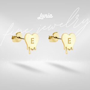May include: A pair of gold heart-shaped stud earrings. Each earring has the letter "E" and a dripping design. The earrings are set against a light grey background with the text "Lunia fine jewelry".