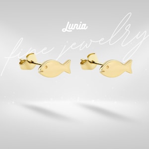 Pode incluir: Um par de brincos de pressão dourados em forma de peixe. Os brincos têm um design simples e minimalista com um pequeno ponto representando o olho. O texto "Lunia fine jewelry" é visível em um fundo cinza claro.