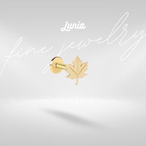 May include: A gold-toned flat back stud earring featuring a maple leaf design. The jewelry piece includes a circular base, a straight post, and a detailed leaf-shaped front. The text Lunia fine jewelry is visible in the background of the image.