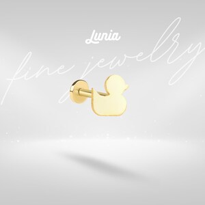 Puede incluir: Un pendiente de botón dorado con forma de pato. El pendiente tiene un poste y una parte trasera plana y circular. La palabra "Lunia" está escrita en cursiva encima del pendiente. Las palabras "fine jewelry" están escritas en cursiva en el fondo.