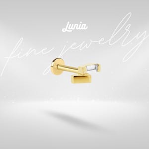 Pode incluir: Brinco dourado com uma pedra preciosa retangular e transparente. O brinco tem uma parte traseira plana e um pino para perfurar. A palavra "Lunia" está escrita em itálico na parte superior da imagem. As palavras "fine jewelry" estão escritas em itálico.