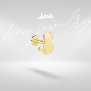 May include: A gold-colored cat-shaped stud earring. The earring features a flat disc backing with a post and a cat silhouette. The word "Lunia" is above the earring, and the words "fine jewelry" are written in cursive.