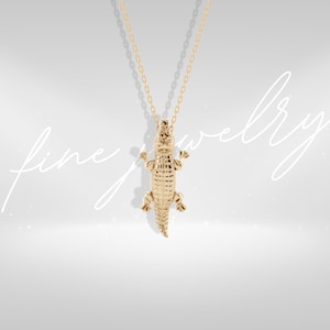Peut inclure: Un collier doré avec un pendentif en forme d'alligator. Le pendentif, suspendu à une fine chaîne, est une représentation détaillée d'un alligator, avec des écailles texturées. Le fond est de couleur neutre. Bijou.