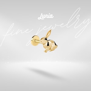 Puede incluir: Un pendiente de oro con un diseño geométrico de conejo. El pendiente tiene una parte posterior plana y un poste para la inserción. La palabra "Lunia" está encima del pendiente, con las palabras "fine jewelry" en cursiva.