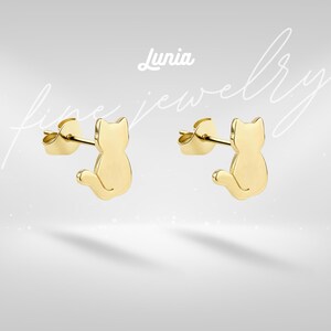 May include: Gold-coloured cat-shaped stud earrings. The earrings are in the shape of a sitting cat with a curved tail. The earrings are displayed on a light grey background with the word "Lunia" and the words "fine jewellery" in a script font.