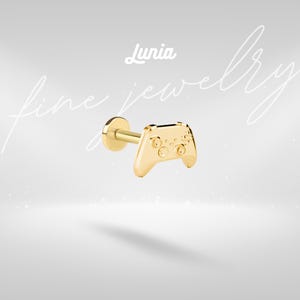 Puede incluir: Un pendiente de botón dorado con forma de mando de videojuego. El pendiente tiene un poste y una parte trasera plana. El diseño del mando incluye botones y una cruceta. La palabra "Lunia" está escrita en cursiva encima del pendiente. Las palabras "fine jewelry" están escritas en cursiva.