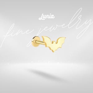 May include: A gold-colored bat-shaped earring stud. The earring has a flat back and a post for piercing. The word "Lunia" is written in cursive above the earring. The words "fine jewelry" are written in cursive in the background.