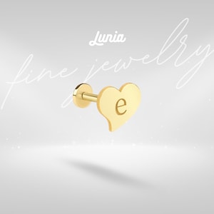 May include: A gold-colored heart-shaped earring with the letter "e" engraved on it. The earring is part of the "Lunia fine jewelry" collection. The earring has a post and a flat back.