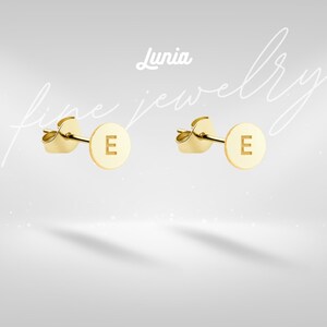 May include: A pair of gold-colored stud earrings. Each earring features a circular disc with the letter "E" engraved in the center. The earrings are set against a soft, light gray background with the words "Lunia fine jewelry" in the image.