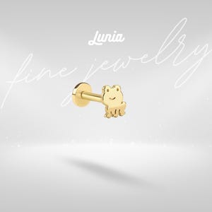 May include: A gold-colored frog-shaped earring with a flat back and a post. The earring has a smiling frog design. The word "Lunia" is written in cursive at the top of the image. The words "fine jewelry" are written in cursive on the left and right sides of the image.