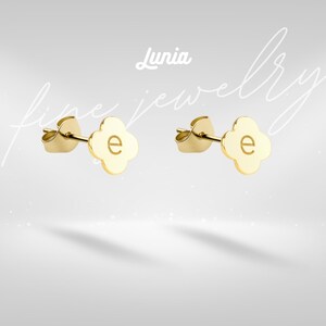 May include: A pair of gold stud earrings, each featuring a four-leaf clover shape with a lowercase letter e engraved in the centre. The earrings include standard post backs and are displayed against a background with the text Lunia fine jewellery.