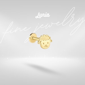 May include: A gold-colored stud earring featuring a sheep design. The earring has a flat back and a post for insertion. The sheep's face is detailed with a smiling expression and a fluffy wool texture. The word "Lunia" is written above the earring.