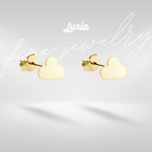 May include: A pair of gold-colored cloud-shaped stud earrings. The earrings are displayed on a light gray background with the word "Lunia" and the words "fine jewelry" in a cursive font.