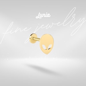 May include: A gold-colored stud earring with an alien face design. The earring has a flat, oval-shaped face with cut-out eyes and a stem for insertion. The word "Lunia" is above the earring. The words "fine jewelry" are written in cursive.