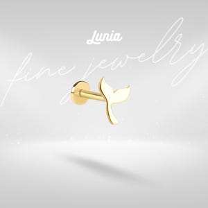 May include: A gold stud earring featuring a whale tail design. The earring has a flat, circular back and a straight post. The whale tail is the main feature, with a curved, symmetrical shape. The word "Lunia" is above the earring.