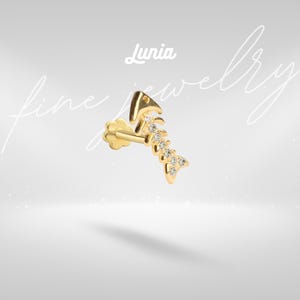Pode incluir: Um brinco dourado em forma de espinha de peixe, com pequenas pedras transparentes incrustadas ao longo da coluna. O brinco inclui um fecho plano em forma de flor e um pino. O texto 'Lunia fine jewelry' é visível no fundo da imagem.