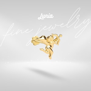 May include: A gold-colored earring featuring a detailed Cupid design with a bow and arrow. The earring is in the shape of a small angel, with the brand name "Lunia" in the upper center. The words "fine jewelry" are written in cursive.