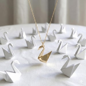 May include: A gold swan pendant necklace with a fine chain. The pendant is a geometric origami design. Several white origami swans surround the necklace, creating a minimalist and artistic display.