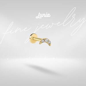 Pode incluir: Um brinco dourado com um design curvo, apresentando três pedras preciosas claras e brilhantes. O brinco tem uma parte traseira plana e circular e um pino para perfuração. A palavra "Lunia" está escrita em itálico acima.