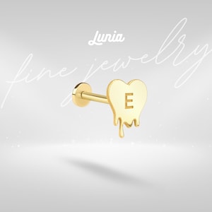 May include: A gold-colored earring with a heart-shaped design and the letter "E" in the center. The heart has a melting effect at the bottom. The earring is from the "Lunia fine jewelry" collection.