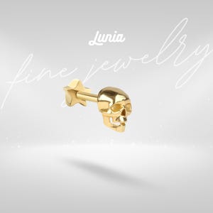 May include: A gold-colored stud earring featuring a three-dimensional skull design on the front and a star-shaped backing. The text Lunia fine jewelry is visible in the background. The piece has a polished metallic finish and a straight post.