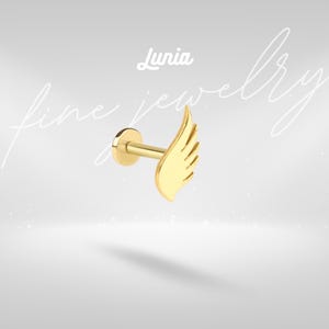 Puede incluir: Un pendiente de color dorado con un diseño de ala. El pendiente tiene una base plana y circular y un poste para la inserción. El ala está detallada con varias líneas. La palabra "Lunia" está encima del pendiente, y las palabras "fine jewelry" están escritas en cursiva.