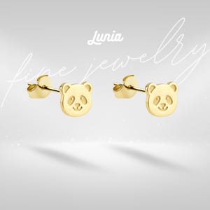 May include: A pair of gold-colored panda bear stud earrings. The earrings have a smooth, polished finish and a post backing. The image includes the text "Lunia fine jewelry" in a script font.