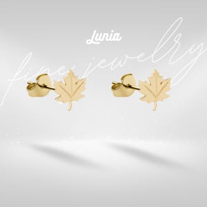 May include: A pair of gold stud earrings shaped like maple leaves with post backs. The text Lunia fine jewellery is displayed in white script above the earrings. The earrings feature a detailed leaf vein pattern and a polished metallic finish.