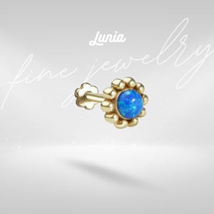 May include: A gold stud earring featuring a round, iridescent blue opal cabochon center surrounded by a scalloped, beaded gold frame. The post has a flower-shaped back. The text 'Lunia fine jewelry' is visible in white script above the earring.
