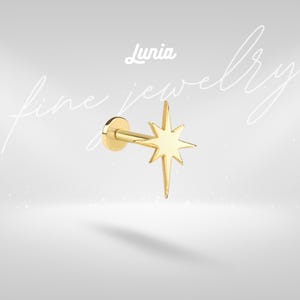 Puede incluir: Un pendiente dorado en forma de estrella con una parte posterior plana y un poste. El pendiente tiene un diseño de estrella con múltiples puntas. La marca "Lunia" está escrita en cursiva encima del pendiente. Las palabras "fine jewelry" están escritas en cursiva.