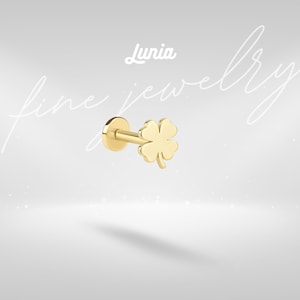 May include: A gold-colored flat back stud earring featuring a four-leaf clover design. The jewelry piece includes a circular base and a post, with the text Lunia fine jewelry displayed in white script and print lettering above the stud.