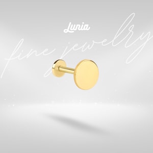 Pode incluir: Um brinco de disco plano dourado com um pino. O brinco tem um design simples e minimalista. A palavra "Lunia" está escrita em itálico acima do brinco. As palavras "fine jewelry" estão escritas em itálico no fundo.