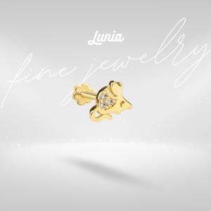 May include: A gold-colored earring in the shape of a mouse, with three small, clear gemstones set in the mouse's body. The earring has a flower-shaped backing and the word "Lunia" is written above the earring.
