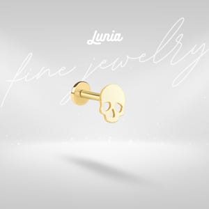 May include: A gold-coloured skull-shaped stud earring. The earring has a flat back and a post for piercing. The word "Lunia" is written in cursive above the earring. The words "fine jewellery" are written in cursive in the background.