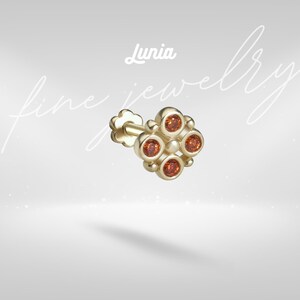 Puede incluir: Un pendiente de botón de oro con un diseño de flor de cuatro pétalos, con cuatro piedras redondas de color naranja engastadas en cada pétalo y una pequeña cuenta central de oro. El texto Lunia fine jewelry aparece sobre el pendiente.