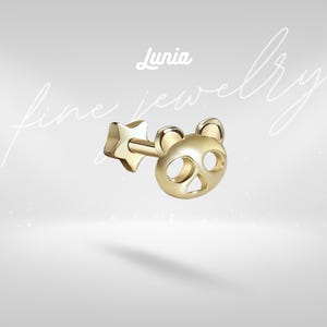 May include: A gold-toned stud earring featuring a panda face design with cut-out eyes and nose, attached to a post with a star-shaped backing. The text Lunia fine jewelry is visible in the background, written in a white script and sans-serif font.