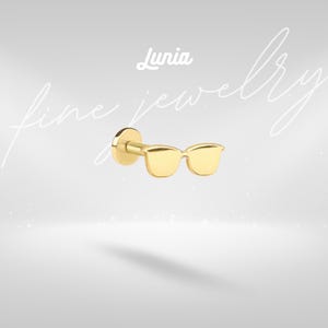 May include: A gold-colored stud earring shaped like sunglasses. The earring has a flat back and a post for insertion. The word "Lunia" is written in cursive above the earring. The words "fine jewelry" are written in cursive in the background.