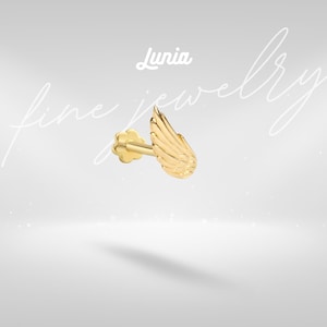May include: A gold-colored earring with a wing design. The earring features a small, flower-shaped backing and a detailed wing shape. The word "Lunia" is written in cursive above the earring. The words "fine jewelry" are written in cursive in the background.