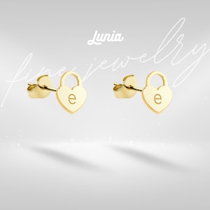 Pode incluir: Um par de brincos de botão dourados em forma de coração. Cada brinco apresenta um design de cadeado com a letra "e" gravada no coração. Os brincos estão sobre um fundo cinza claro com as palavras "Lunia fine jewelry".