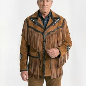Peut inclure: Veste en daim marron avec des franges, des bordures noires et des coutures décoratives. La veste a une fermeture boutonnée et est portée avec un pantalon marron. La veste comporte des franges sur la poitrine, les manches et le dos.
