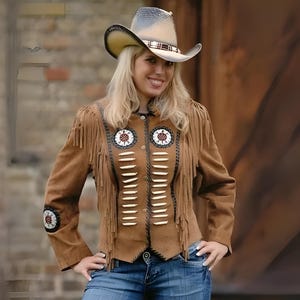 May include: A tan suede jacket featuring long fringe detailing on the shoulders and chest, white bone-like accents, and circular beaded patches on the chest and sleeves. The jacket is paired with a light-colored cowboy hat with a dark band.